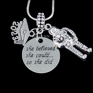 May include: Silver necklace with a graduation cap charm, an astronaut charm, and a round charm with the text "she believed she could... so she did".
