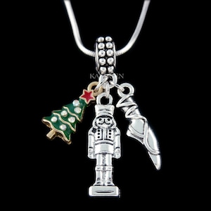May include: Silver necklace with a charm featuring a nutcracker, a Christmas tree, and a ballet slipper. The charms are silver with red, green, and gold accents.