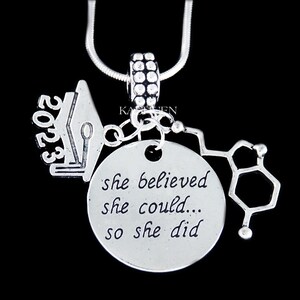 May include: Silver necklace with a graduation cap charm, a charm with the text "she believed she could... so she did", and a charm shaped like a chemical structure. The graduation cap charm has the year 2023 on it.