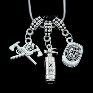 May include: Silver-coloured necklace with firefighter charms, including crossed axes, a fire extinguisher, and a helmet. The necklace has the text "FD" on the axe charm and "FIRE" on the extinguisher. The background is black.