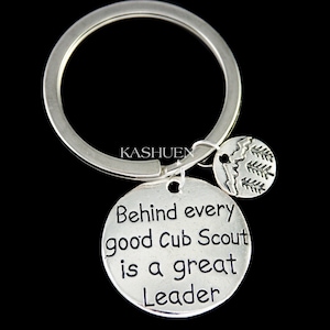 May include: Silver keychain with a round charm that says "Behind every good Cub Scout is a great Leader". The charm has a smaller charm attached to it with a design of pine trees.
