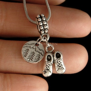 May include: Silver necklace with a charm of a pair of hiking boots and a round charm with a mountain scene and pine trees.