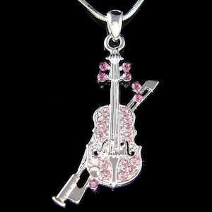 Puede incluir: Un collar con un colgante de violín plateado con detalles de cristal rosa. El violín está colocado sobre un arco plateado.