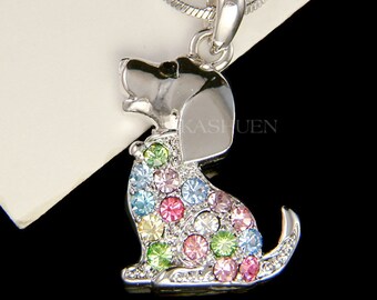 Può includere: Collana d'argento con un ciondolo a forma di cucciolo tempestato di cristalli. Il cucciolo è seduto con la testa inclinata e ha un occhio nero. I cristalli sono multicolori, tra cui rosa, blu, verde e giallo.