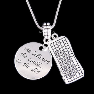 Könnte beinhalten: Silberne Halskette mit zwei Anhängern. Ein Anhänger ist ein Kreis mit dem Text "she believed she could... so she did". Der andere Anhänger ist eine Tastatur mit den in alphabetischer Reihenfolge beschrifteten Tasten.