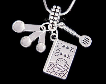 May include: Silver necklace with a charm featuring a cookbook, measuring spoons, and a spatula. The cookbook charm has the words "Cook Book" on the cover.