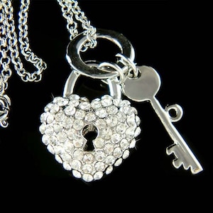 以下が含まれることがあります： クリアクリスタルをちりばめたハート型のペンダントが付いたシルバーカラーのネックレス。小さな鍵のチャームがハート型のロックの隣にぶら下がっています。ネックレスチェーンは画像の左側に表示されています。