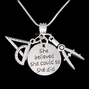 May include: Silver necklace with a round charm that reads "She believed she could so she did". The charm is attached to a silver protractor, compass, and set square.