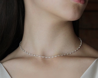 May include: A delicate silver chain necklace with a simple design. The necklace is worn by a person with dark hair and is shown against a wooden background.