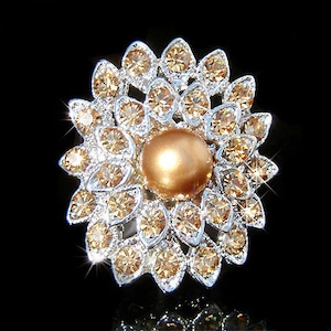 Puede incluir: Un broche de plata en forma de flor con una perla grande, redonda y marrón claro en el centro. La flor está hecha de muchas pequeñas piedras brillantes transparentes.