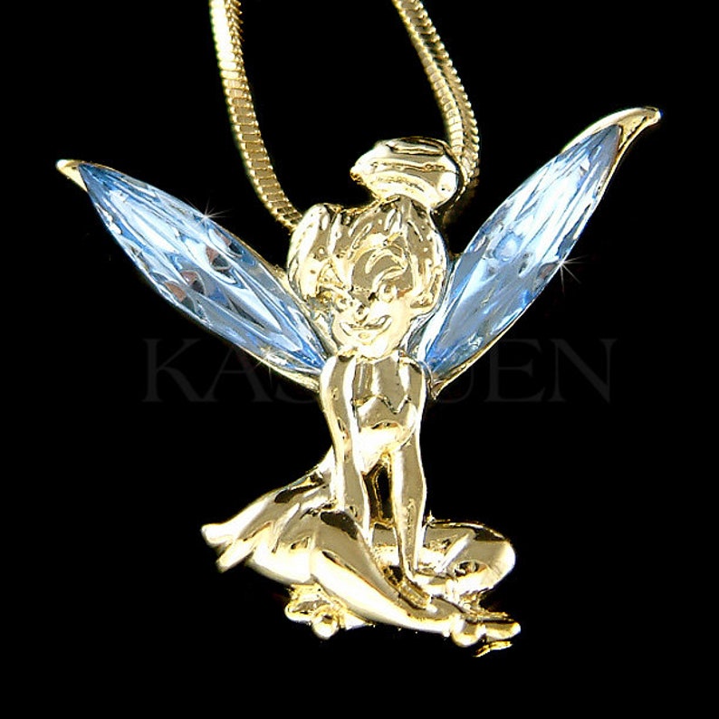 Puede incluir: Colgante de hada dorado con alas azul claro, suspendido de una cadena dorada. El hada est&aacute; sentada con los brazos cruzados. El colgante tiene la palabra "KASDEN" impresa.
