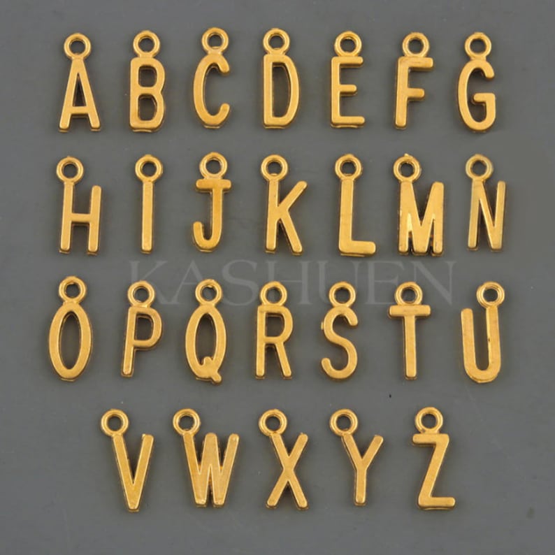 Puede incluir: Abalorios dorados con letras del alfabeto, cada uno con un peque&ntilde;o lazo en la parte superior para sujetarlos a joyas u otras manualidades. Las letras de la A a la Z se muestran en una cuadr&iacute;cula ordenada sobre un fondo gris. Los abalorios son todos en una fuente sans-serif.