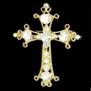 May include: A gold-tone cross brooch with clear rhinestones. The cross is ornate with a detailed design and has a pin on the back.