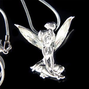 Puede incluir: Collar con colgante plateado que representa un hada. El hada est&aacute; representada con alas detalladas y est&aacute; sentada. La cadena del collar es una fina cadena de serpiente plateada. El fondo es negro.