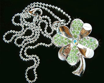 Puede incluir: Un collar de plata con un colgante de trébol de cuatro hojas. El trébol está hecho de plata y tiene cristales verdes en cada hoja. El collar está sobre un fondo negro.