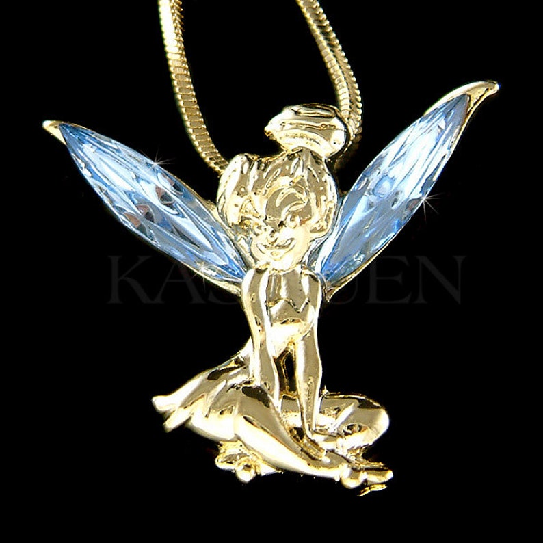 K&ouml;nnte beinhalten: Eine goldfarbene Halskette mit einem Anh&auml;nger, der eine sitzende Fee mit hellblauen Kristallfl&uuml;geln zeigt. Die Fee hat ein zartes, detailliertes Design und einen s&uuml;&szlig;en Ausdruck.