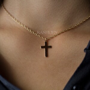 May include: A gold-colored necklace with a cross pendant. The cross is a simple, classic design, suspended from a delicate chain. The necklace is worn against a dark background, highlighting the jewelry's shine. The brand name "KASHUEN" is visible.