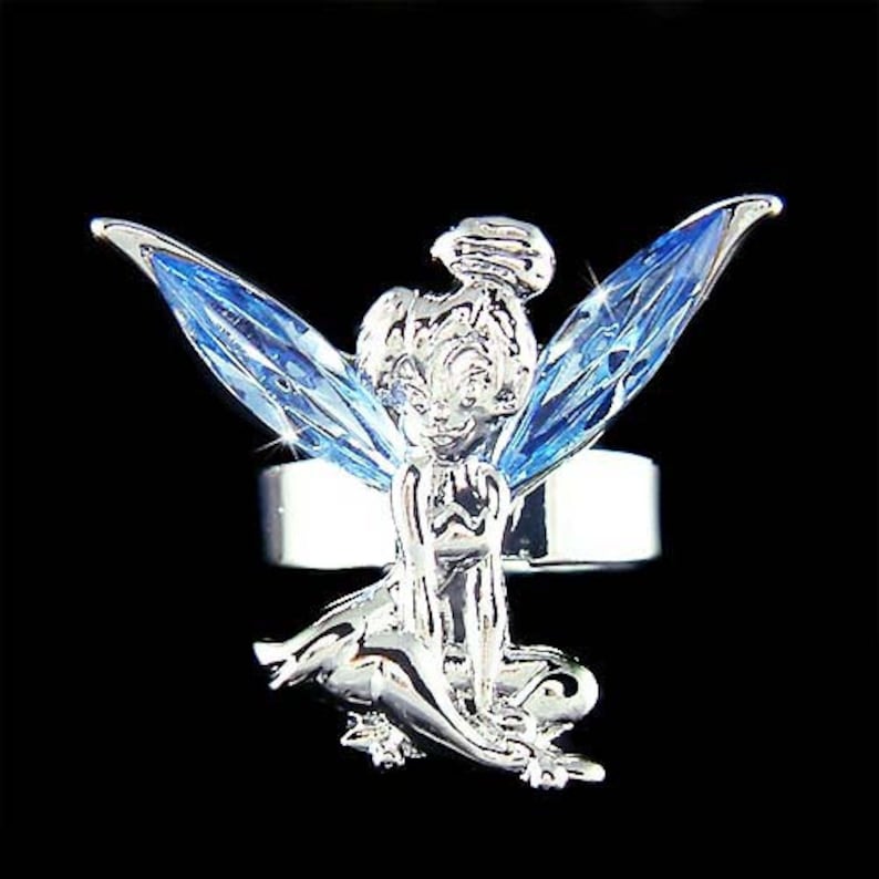 May include: A silver ring featuring a blue crystal fairy design. The fairy has wings and is perched on a ring band.