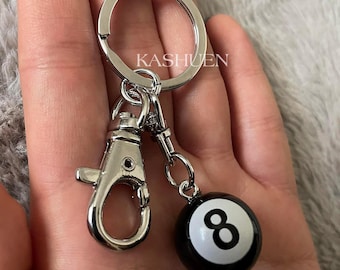 May include: Silver-toned keychain with a lobster clasp and a round key ring. A small black and white eight ball charm is attached. The text "KASHUEN" is printed on the key ring.