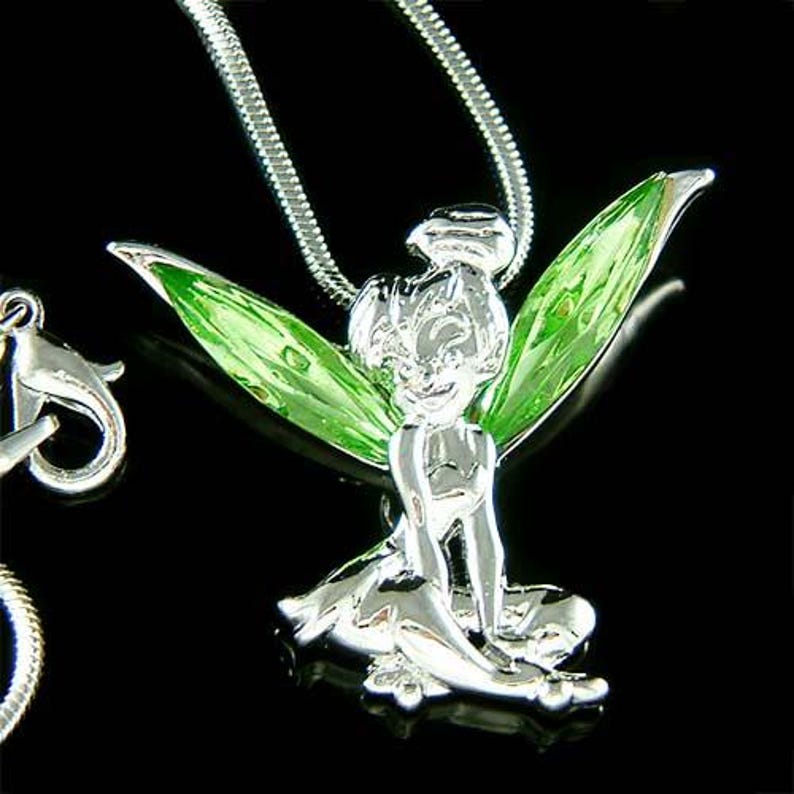 Puede incluir: Un collar con colgante plateado que representa un hada con alas verdes. El hada est&aacute; sentada, con una expresi&oacute;n sonriente. El collar tiene una cadena plateada y un cierre de mosquet&oacute;n.