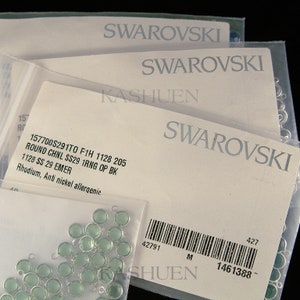 Peut inclure: Gros plan sur un paquet de perles de cristal vert clair avec des boucles argent&eacute;es. L'emballage est &eacute;tiquet&eacute; avec le texte "Swarovski" et le code produit "157700S291T0 F1H 1128 205".