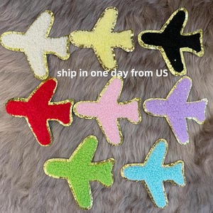 May include: A collection of colorful airplane patches in white, yellow, black, red, pink, purple, green, and blue. Each patch has a gold glitter border and a textured, fuzzy appearance. The patches are arranged on a gray, furry surface.