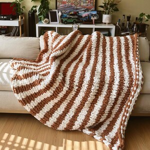 May include: A chunky knit blanket in a brown and cream striped pattern is draped over a beige sofa. The blanket's thick yarn creates a textured, cozy appearance. The room has a warm, inviting atmosphere with decorative items on shelves.