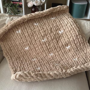 May include: A chunky knit blanket in a light brown color, adorned with small white heart-shaped accents. The blanket is folded over, showcasing its thick, textured yarn. It is resting on a neutral-toned surface, suggesting a cozy home decor item.