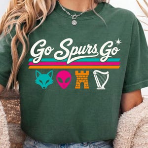 May include: A dark green short-sleeve t-shirt featuring the white text Go Spurs Go above three horizontal stripes in teal, pink, and orange. Below the stripes are four icons: a teal fox head, a pink alien head, an orange castle, and a white harp.