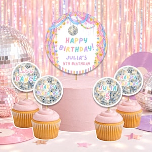 Peut inclure: Un ensemble de décorations pour gâteaux et cupcakes comprenant une grande pancarte circulaire avec le texte Happy Birthday Julia's 5th Birthday, entourée de quatre petits disques illustrés de boules à facettes et du texte Let's Dance et Julia 5.