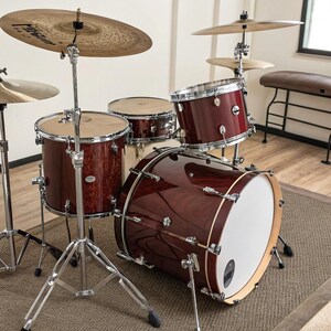 May include: A complete drum kit with a deep red wood grain finish. The kit includes a bass drum, snare drum, and multiple tom-toms, along with cymbals and stands. The drum kit is set up on a brown rug.