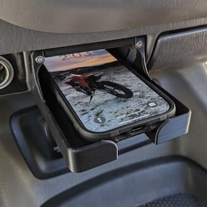 May include: A black phone holder with a phone inside. The phone is in a black case and has a screen displaying a photo of a dirt bike. The phone holder is mounted in a car's centre console.