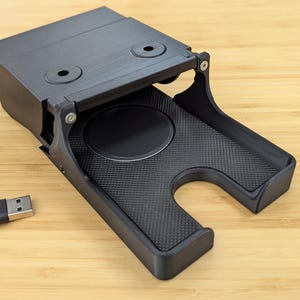 May include: A black plastic charging stand with a built-in wireless charging pad and a USB cable. The stand has a cutout for a phone and a textured surface.