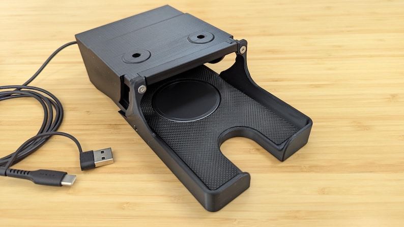 May include: A black plastic charging stand with a built-in wireless charging pad and a USB cable. The stand has a cutout for a phone and a textured surface.