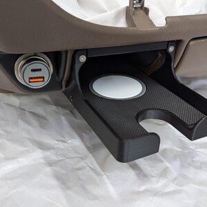 May include: A black plastic cup holder with a gray rubber insert and a black textured surface. The cup holder is attached to a brown surface and has a small black power outlet with two USB ports.