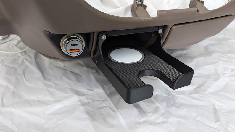 May include: A black plastic cup holder with a gray rubber insert and a black textured surface. The cup holder is attached to a brown surface and has a small black power outlet with two USB ports.