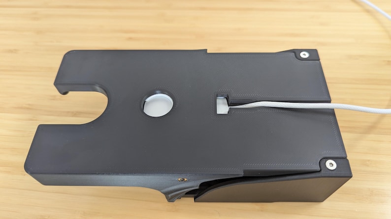 May include: A black plastic cable management box with a white cable running through it. The box has a circular opening and a slot for the cable.