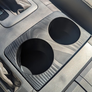 Puede incluir: Inserto personalizado para la consola central de un vehículo con dos portavasos. El inserto es de material gris oscuro con un patrón de vetas de madera. Los portavasos son negros y redondos.