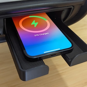 Puede incluir: Un teléfono negro con una animación de carga en la pantalla se coloca en una plataforma de carga inalámbrica negra. La pantalla del teléfono muestra "21% cargado".
