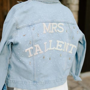 Op de afbeelding: Lichtblauwe spijkerjas met de woorden "MRS TALLENT" in het wit, versierd met parels en kleine gouden accenten. De jas heeft een klassieke stijl, perfect voor een bruiloft of bruidsevenement.