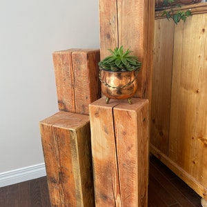 May include: Three rectangular wooden pillars of varying heights arranged in a tiered formation. The wood features a natural grain with visible cracks and a light brown finish. A small copper-coloured pot with a succulent sits on the middle pillar.