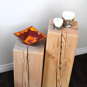 May include: Two wooden end tables with a natural wood finish. The table on the left has a red and orange ceramic bowl on top. The table on the right has two white candles on top.