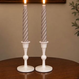May include: Two lit taper candles with a grey, twisted, spiral design. Each candle is held in a smooth, white, pedestal-style candle holder with a round base, a slender stem featuring a central spherical accent, and a flared top cup.