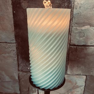 May include: A large, teal-colored pillar candle with a spiral design, resting on a black metal candle holder. A small, gold-colored heart-shaped decoration sits atop the candle. The candle is lit, emitting a warm glow.