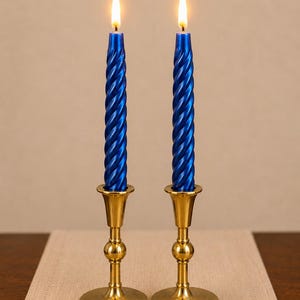May include: Two metallic blue spiral taper candles with lit flames, each standing upright in a polished gold-toned metal candlestick holder. The candles feature a twisted, ribbed texture and are positioned side-by-side on a flat surface.