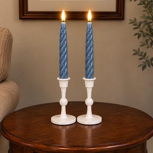 May include: Two blue spiral taper candles with glowing faux flames, each set in a white candlestick holder featuring a round base, a slender stem with a central spherical accent, and a flared top cup for holding the candle securely.