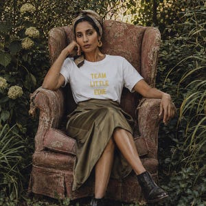 South Asian woman wearing a white Team Little Edie graphic T-shirt, olive satin skirt and headscarf, seated in a faded floral armchair surrounded by an overgrown garden.