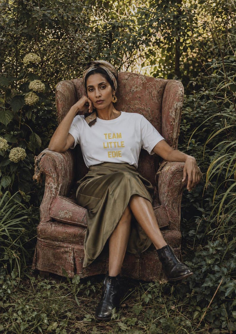 South Asian woman wearing a white Team Little Edie graphic T-shirt, olive satin skirt and headscarf, seated in a faded floral armchair surrounded by an overgrown garden.