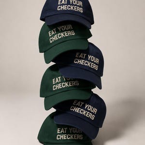Stack of navy and forest-green dad caps embroidered with the phrase Eat Your Checkers in cream lettering.