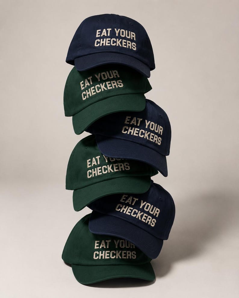 Stack of navy and forest-green dad caps embroidered with the phrase Eat Your Checkers in cream lettering.